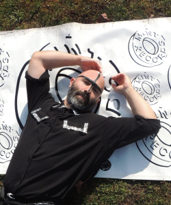 the artist devours wearing a black shirt and laying down on a mint records banner 